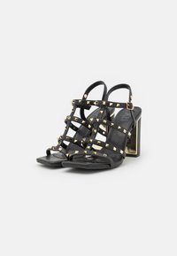 Sandalias de tacón negro con un diseño de tiras que presenta tachuelas de pirámide doradas, punta cuadrada y un tacón cuadrado con acentos metálicos.