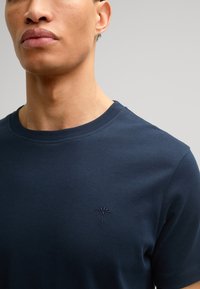 Granatowa koszulka okrągłym dekoltem, wykonana z miękkiej bawełny. Posiada subtelny haftowany wzór na lewej piersi. Gładka tekstura, krótkie rękawy.