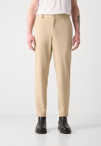 Beige teksturerte bukser med knappelukking og sidelommer, kombinert med svarte ankelstøvler foran en enkel bakgrunn.