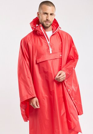 Homme portant un poncho de pluie imperméable rouge vif avec capuche et demi-fermeture éclair à l'avant, debout devant un fond clair uni.