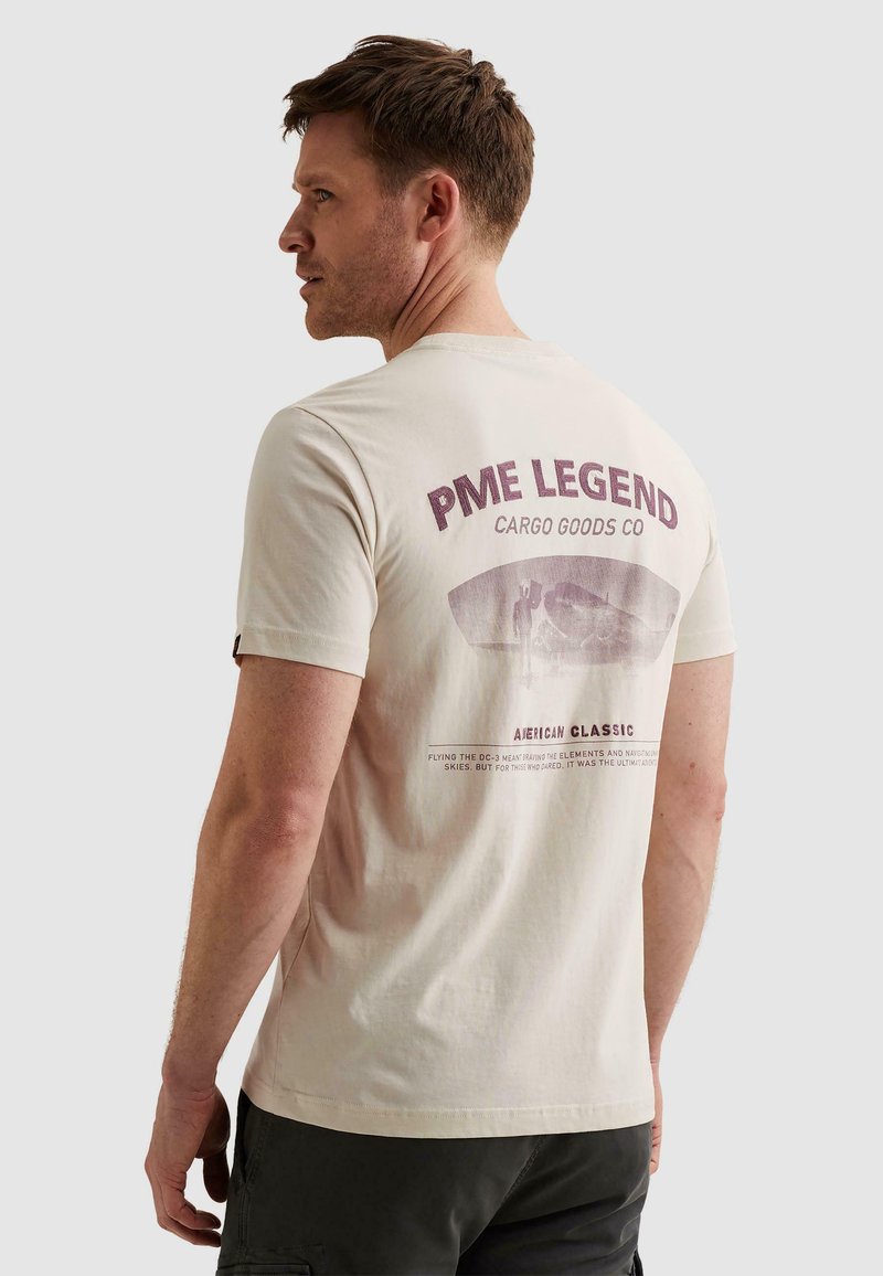 Homme portant un t-shirt PME Legend écru avec un short cargo, légèrement tourné pour montrer le graphisme de la marque et le texte au dos.