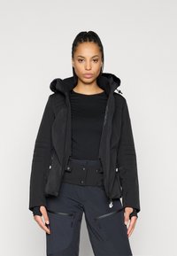 Μαύρο σακάκι σκι με κουκούλα επενδεδυμένη με γούνα, κλείσιμο με φερμουάρ, υφή υφάσματος και ραμμένες λεπτομέρειες στα μανίκια. Φορεμένο πάνω από μαύρο βασικό στρώμα και παντελόνι.