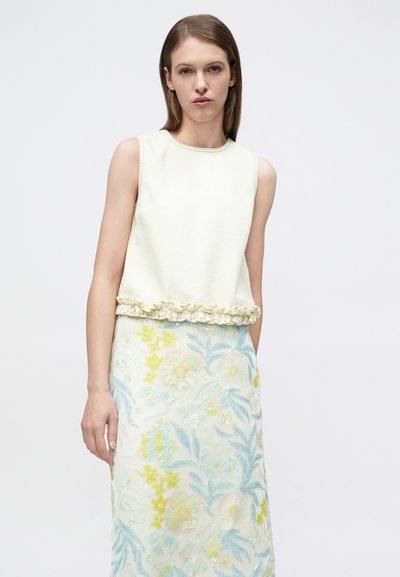 Femme portant un haut crème sans manches avec ourlet à volants et une jupe brodée de fleurs aux tons bleu, jaune et blanc sur fond blanc.