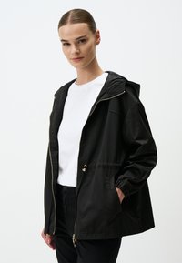 Zwarte hoodie gemaakt van gladde stof, met een ritssluiting, elastische manchetten en een verstelbare taille. Draagt over een wit t-shirt.