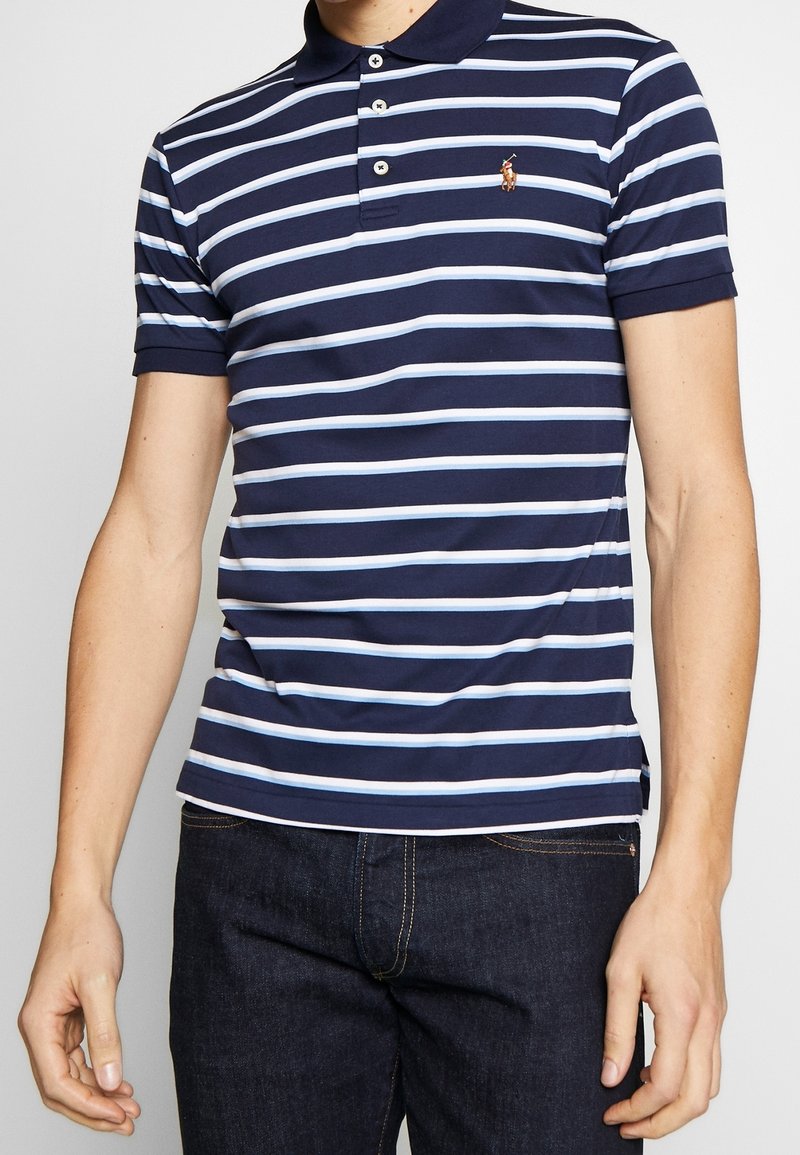 Námořnická modrá polo košile s světle modrými pruhy, krátké rukávy, zapínání na tři knoflíky a malá vyšívaná značka na hrudi.