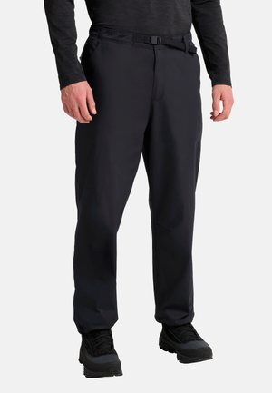 Mann trägt schwarze Freizeithose mit verstellbarem Gürtel, schwarzes langärmeliges Oberteil und schwarze Outdoorschuhe vor weißem Hintergrund.