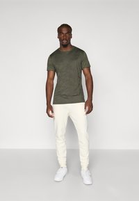 Olive green short-sleeve t-shirt with a subtle pattern, paired with cream-colored joggers and white sneakers on a plain background.