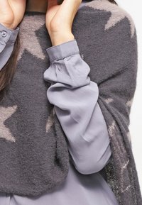 Châle en tricot gris avec des motifs d'étoiles drapé sur un haut gris clair. Le châle a une texture douce et des bords frangés, ajoutant du détail.