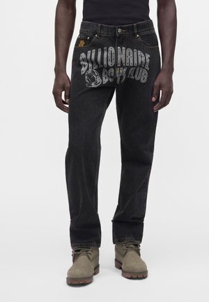 Jean noir avec un grand texte "Billionaire Boys Club" et un graphisme de casque d'astronaute sur le devant, porté avec des bottes beige.