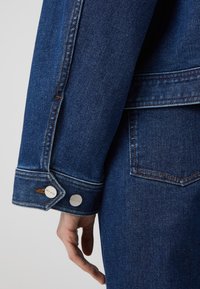 Tummansinisen farkkutakin hihansuu, jossa hopeiset napit ja oranssit kontrastitikkaukset. Sileä pinta ja klassinen muotoilu.