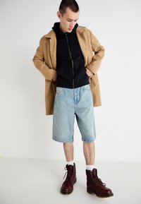 Outfit informale che presenta un cappotto beige, una felpa nera con zip, dei pantaloni shorts in denim azzurro chiaro e stivaletti borgogna con lacci e cuciture gialle.