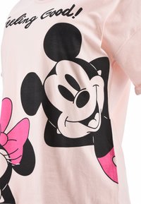 Camiseta de algodón rosa claro con personajes de dibujos animados en negro y rosa, incluyendo un ratón sonriente y un lazo, con el texto "Feeling Good!" en la parte superior.