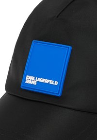Must kaal kork, millel on sile tekstuur, esitleb silmapaistvat sinist ruutpatchi, millel on kõrgendatud valge kiri "KARL LAGERFELD JEANS."
