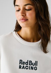 Sudadera blanca con cuello redondo y textura suave, que destaca el texto "Red Bull RACING" en letras negras en negrita.