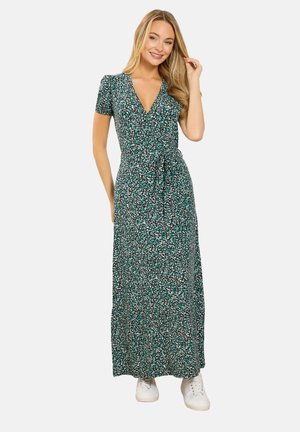 Kvinde smilende iført en kortærmet, V-udskåret, grøn blomster indpakningskjole med taljebånd og hvide sneakers, stående mod en ensfarvet baggrund.