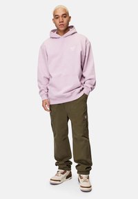 Ljuslila hoodie med ficka fram och logotyp, kombinerad med olivgröna cargobyxor och vita sneakers med bruna detaljer.