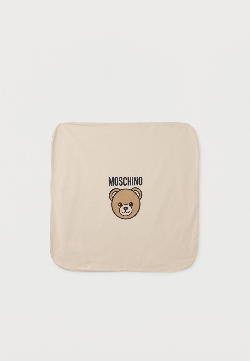 Μπεζ υφασμάτινο τετράγωνο με στρογγυλεμένες γωνίες που απεικονίζει ένα γραφικό αρκούδας και τη λέξη «MOSCHINO» με μαύρα γράμματα πάνω από το πρόσωπο της αρκούδας.
