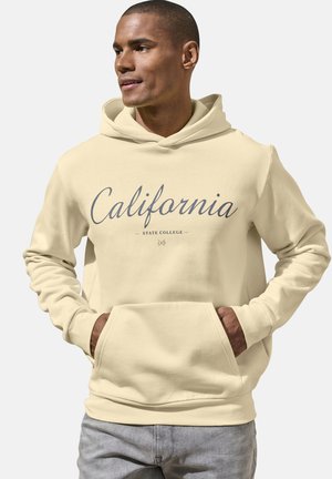 Mann trägt beigen Hoodie mit „California State College“-Aufschrift, Hände in der vorderen Tasche, schaut zur Seite vor einfachem Hintergrund.