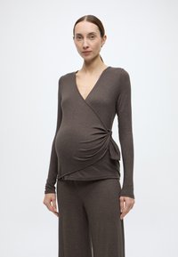 Καφέ τοπ από απαλό, ελαστικό ύφασμα· διαθέτει μακριά μανίκια και λεπτομέρεια με κόμπο στο πλάι. Στυλιζαρισμένο με παντελόνι στο ίδιο χρώμα.