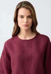 Top in lino bordeaux con scollatura rotonda e maniche ampie e morbide. Il tessuto presenta una sottile texture e una caduta morbida.