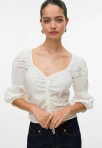 Blusa bianca con scollatura a V, maniche a sbuffo e dettagli arricciati sul davanti; presenta accenti in pizzo sulle maniche; tessuto leggero e testurizzato.