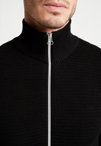 Černá svetr s vysokým žebrovaným límcem a texturovanou tkaninou. Obsahuje stříbrný zip a těsný střih kolem krku.