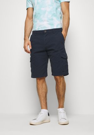 Homme portant un short cargo bleu marine, un t-shirt tie-dye bleu clair, des chaussettes blanches et des baskets blanches, debout avec une main dans la poche.