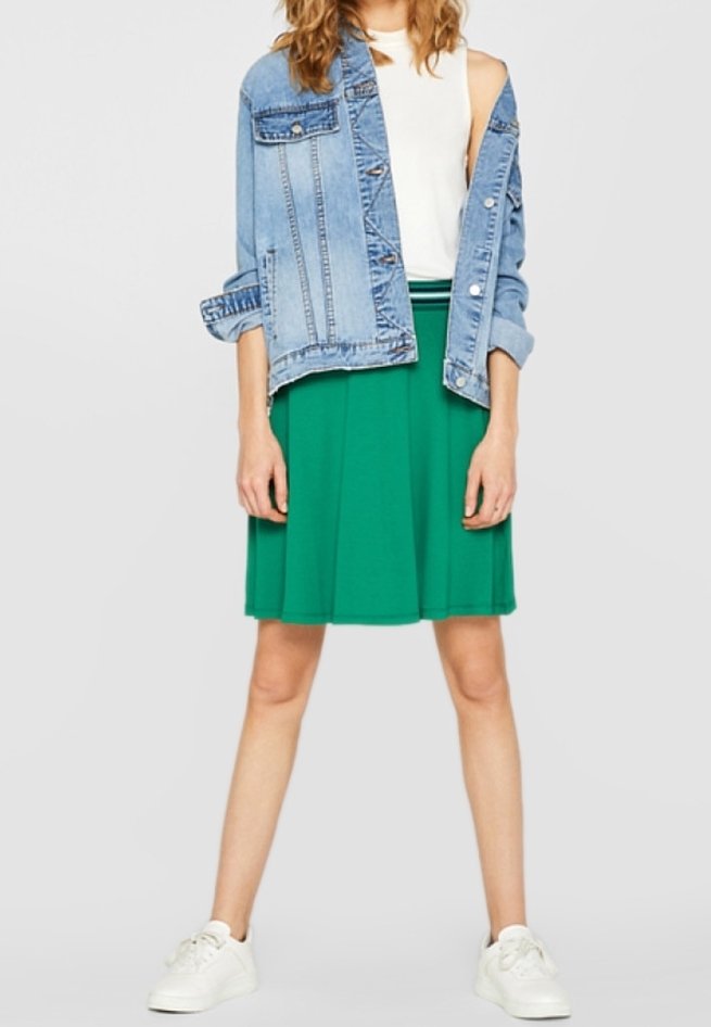 Femme portant une jupe verte, un haut blanc sans manches, une veste en jean bleue partiellement enlevée et des baskets blanches, debout les jambes écartées sur fond blanc.