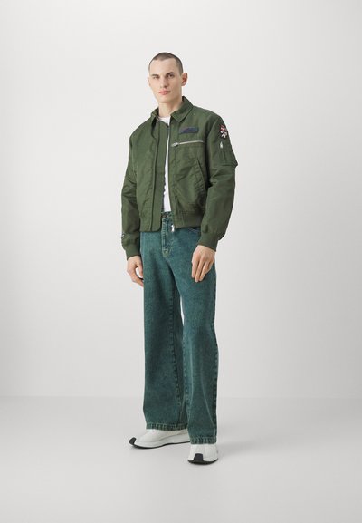 Giacca bomber verde con colletto, zip frontale e toppe ricamate, abbinata a jeans a gamba larga blu e sneakers bianche.