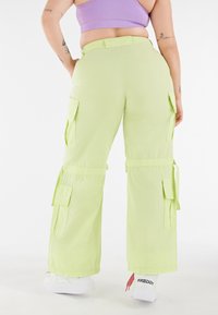 Pantalones cargo en verde claro, con múltiples bolsillos laterales, lazos ajustables en los puños y un ajuste holgado. Combinados con zapatillas blancas.