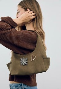 Olivengrøn ruskinds taske med to dekorative mærker: et sølvblomstermotiv og en hjerteformet accent i brun. Funktionen har en bred, buet form.