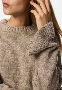 Beige kötött pulóver bordás kerek nyakkal és textúrált ujjakkal. Puha gyapjú anyag világos és sötét szálak keverékét tartalmazza.