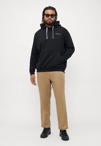 Hanorac negru din bumbac cu un buzunar frontal, șnururi albe și un logo mic. Asortat cu pantaloni bej și adidași negri.