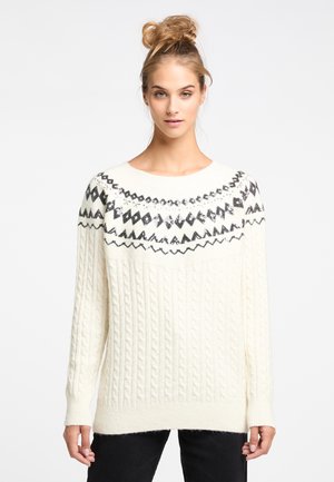 Jonge vrouw met blond haar in een knot, die een witte kabelgebreide trui draagt met zwarte geometrische patronen op het juk en een zwarte broek.