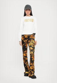 Camicia bianca a maniche lunghe con logo "Versace Jeans Couture", abbinata a pantaloni neri svasati con complessi motivi floreali dorati. Cappello nero e scarpe chunky.