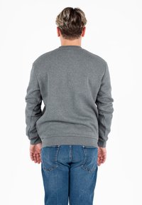 Persona con capelli corti e mossi, che indossa una felpa grigia e jeans blu, in piedi con le spalle rivolte verso di noi su uno sfondo bianco.