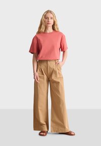 Korallfärgad bomullströja med uppvikt ärm, kombinerad med beiga vida byxor som framhäver en avslappnad passform och subtila veck. Bruna sandaler.