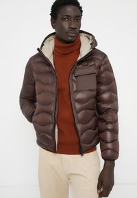 Braune Steppjacke mit horizontalen Baffeln, hellbeigem Futter und Reißverschlusstasche. Getragen über einem rostfarbenen Rollkragenpullover, geschichtet für Wärme.