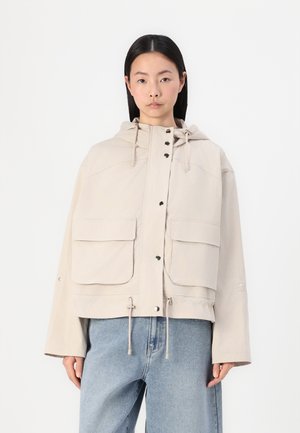 Veste à capuche beige clair avec de grandes poches avant, fermeture à bouton et ourlet à cordon. Fabriquée dans un tissu texturé, elle présente un design minimaliste.