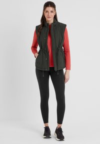 Dark green quilted vest with a high collar and drawstring waist, worn over a red shirt, paired with black leggings and athletic shoes.