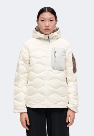 Jeune personne portant une veste matelassée blanche avec capuche et poches zippées contrastées sur la poitrine et les bras, debout devant un fond uni.