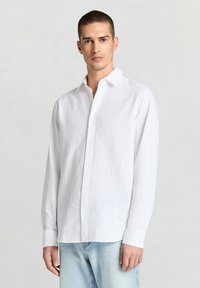 Chemise blanche à manches longues en tissu léger, dotée d'un devant à boutons, d'un col pointu et d'une texture lisse, associée à un jean bleu clair.