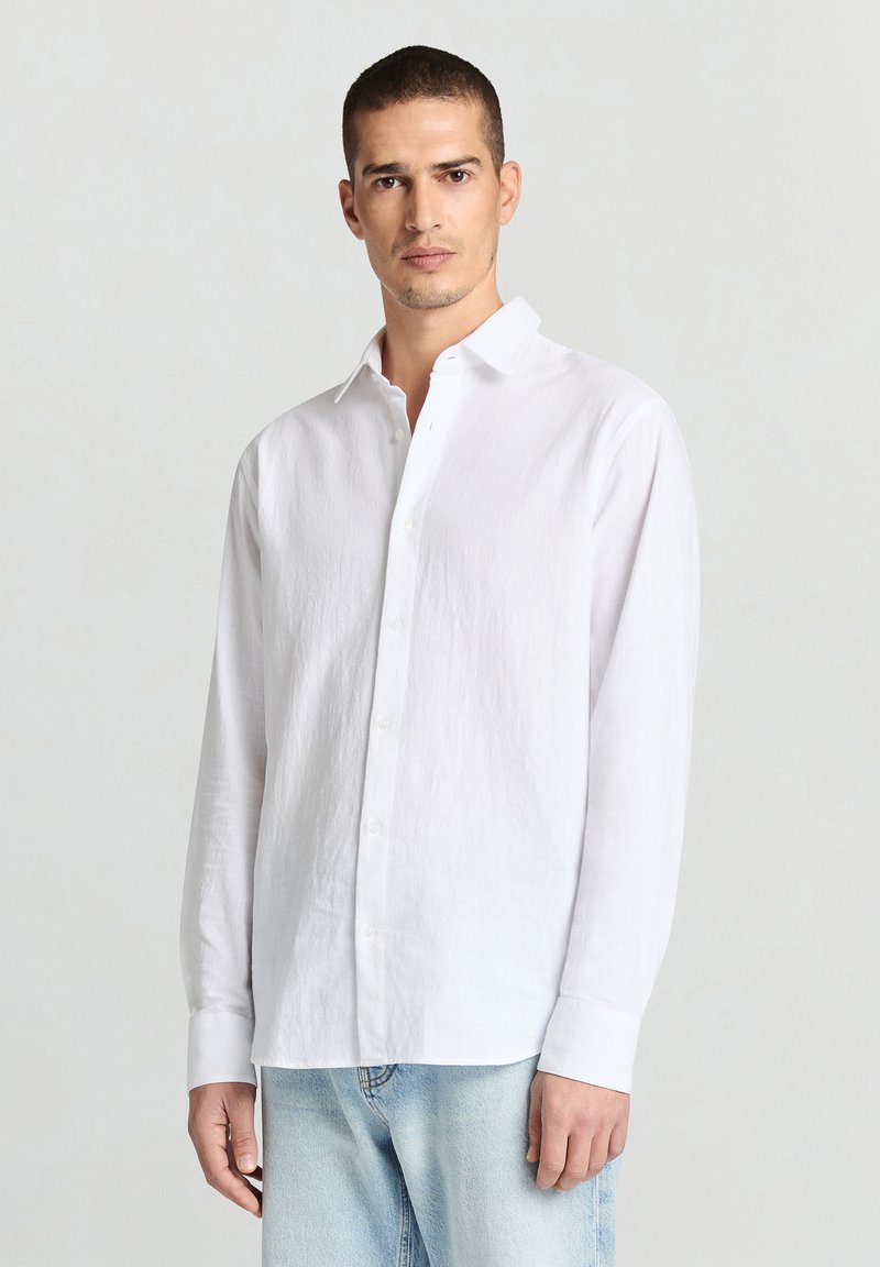 Chemise blanche à manches longues en tissu léger, dotée d'un devant à boutons, d'un col pointu et d'une texture lisse, associée à un jean bleu clair.