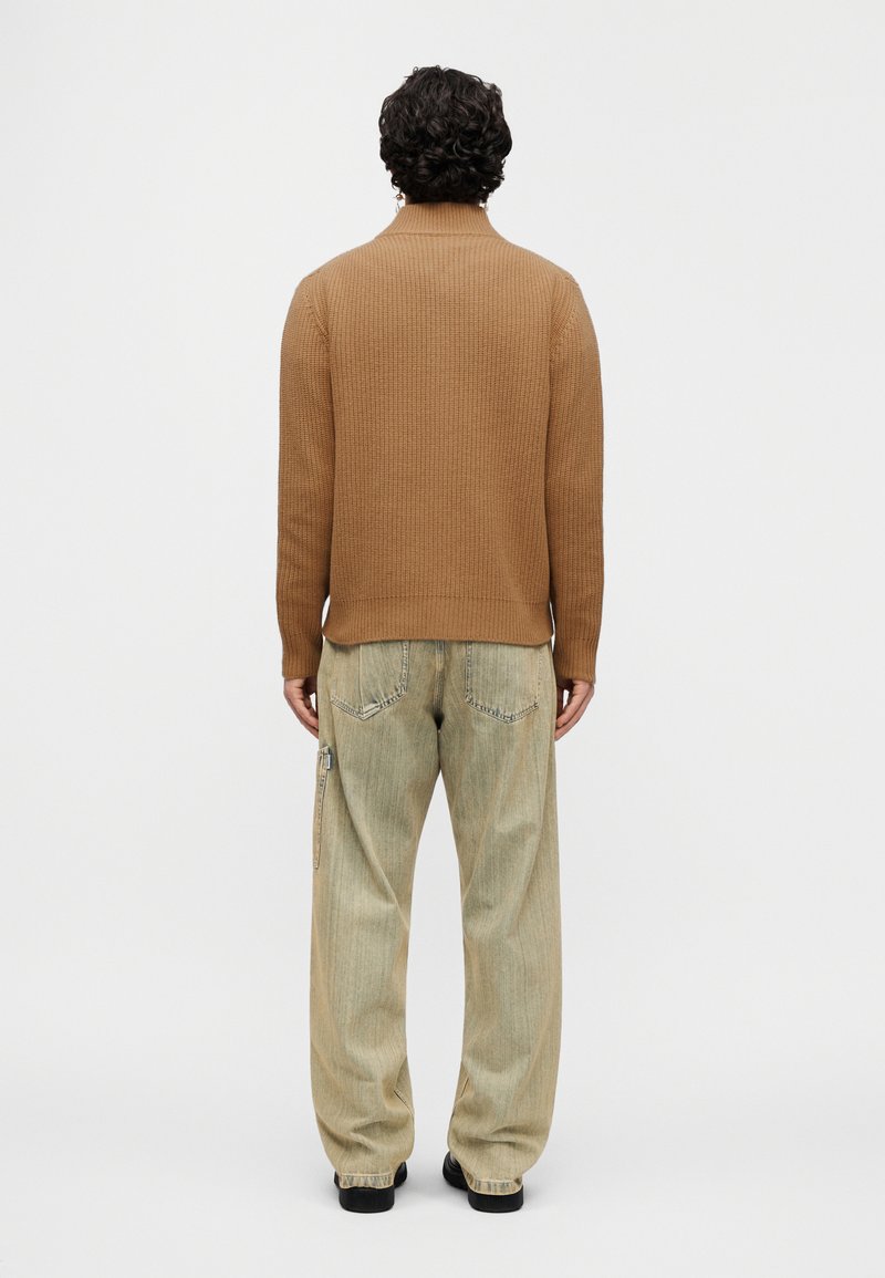 Brown ribbed knitted sweater with a high neck, paired with loose, light denim pants featuring back pockets and a side zipper.