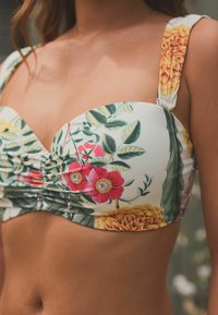 Bikini gornji dio s cvjetnim uzorkom u bijeloj boji sa zelenim listovima, ružičastim i narančastim cvjetovima. Ima nabran središnji dio i široke naramenice s cvjetnim naglascima.