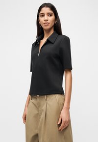 Μαύρο κοντομάνικο πόλο με φερμουάρ μπροστά, κατασκευασμένο από λείο υλικό, με μυτερό γιακά και διακριτική κεντημένη λογότυπο.