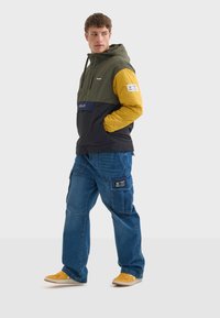 Chaqueta cortavientos a bloques de color con paneles verdes, amarillos y negros; cuenta con capucha, bolsillo frontal y parche con logo. Combinada con vaqueros cargo azules y zapatillas amarillas.