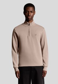 Beige ritspullover gemaakt van zachte stof met een hoge kraag en lange mouwen; voorzien van een klein, geborduurd logo op de borst.