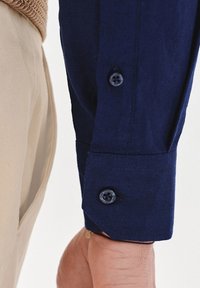 Granatowa koszula z długim rękawem, z zapięciem na guzik, o gładkiej strukturze i jednym ciemnym guziku. Obok widoczny beżowy materiał.