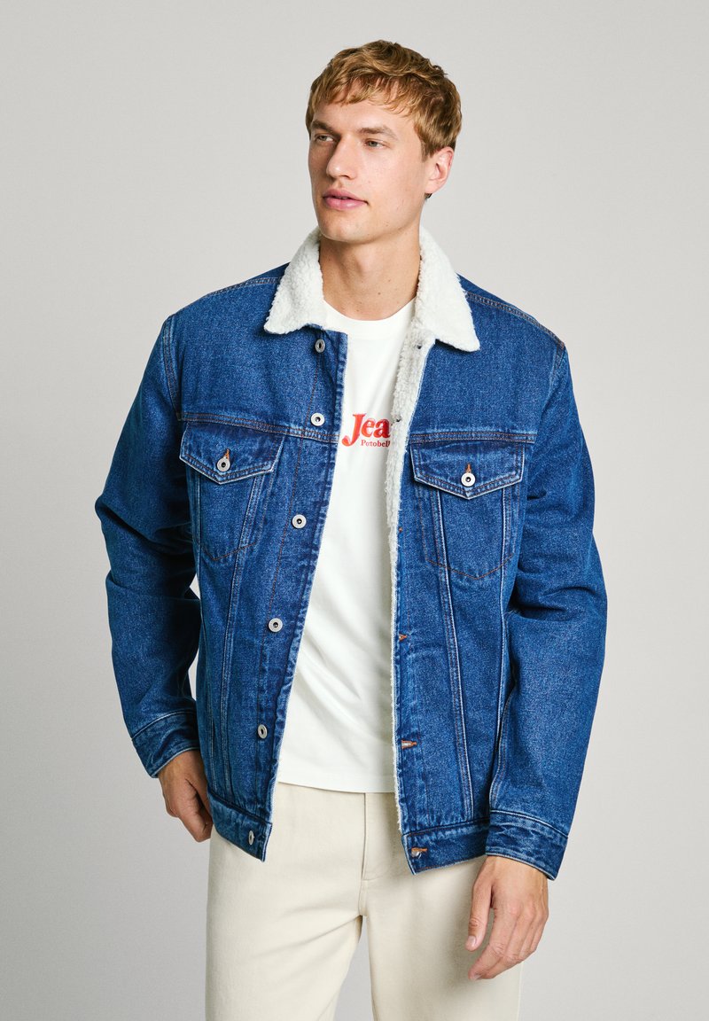 Denim-jakk, millel on karvane kaelus, sügavat sinist värvi, nupulukke, kaks rinna-taskut ja lõtv istuvus, kantud valge T-särgi üle.